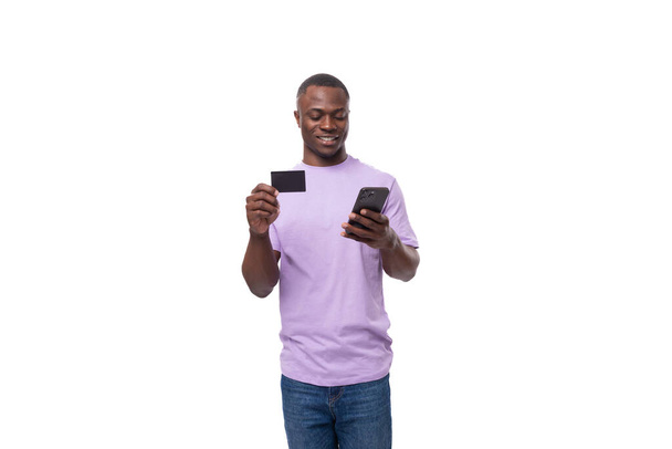 een jonge vrolijke slanke Amerikaanse student man gekleed in een licht lila T-shirt en jeans maakt gebruik van een online smartphone en een creditcard. - Foto, afbeelding