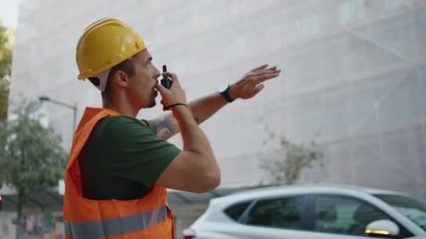 Hardhat-mies antaa ohjeita työntekijöille, jotka käyttävät radiopuhelinta työmaan lähikuvassa. Vakava latinoinsinööri oranssissa liivissä pitäen kannettavaa radiota puhumassa kadulla. Rakennuspäällikkö osoittaa - Materiaali, video