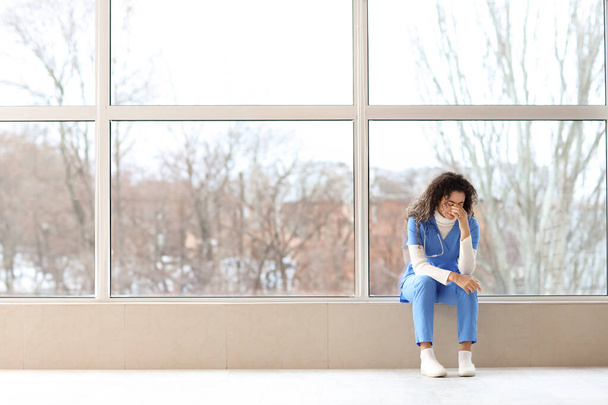 Väsynyt afroamerikkalainen lääketieteen harjoittelija istuu ikkunalaudalla klinikalla - Valokuva, kuva