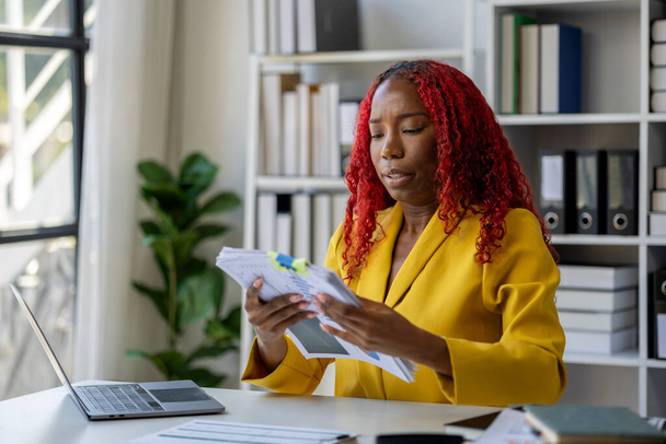 Piękna afrykańska bizneswoman siedzi w prywatnym biurze w firmie, Kobieta pracownik szuka informacji, aby przygotować się do spotkania, Siedząc przy biurku robi różne rzeczy. - Zdjęcie, obraz