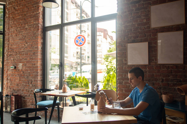 Ευτυχισμένος Καυκάσιος μαθητής Απολαμβάνοντας τον καφέ και την περιήγηση στα μέσα κοινωνικής δικτύωσης σε ένα Sunny Coffee Bar μετά τα μαθήματα - Φωτογραφία, εικόνα