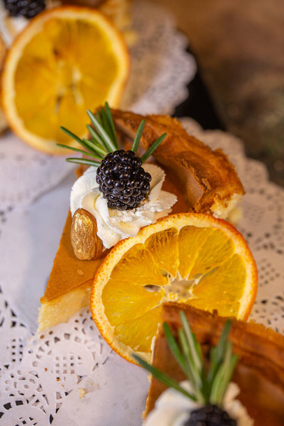 Eine dekadente Präsentation von vier Scheiben cremigem Käsekuchen mit leuchtenden Orangenscheiben und saftigen Brombeeren auf einem zierlichen weißen Deckchen. - Foto, Bild