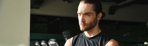 Un hombre musculoso en una camiseta negra de pie con confianza en un gimnasio. - Foto, imagen