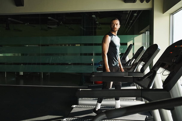 Een atletische man in actieve kleding staat vol vertrouwen op een loopband in een fitnessruimte, focus en vastberadenheid geëtst op zijn gezicht. - Foto, afbeelding