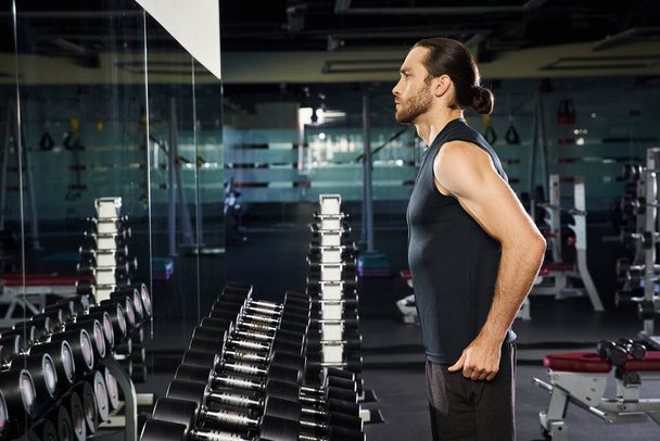 Un homme athlétique en tenue active se tenant debout en toute confiance devant un rack d'haltères, se préparant pour une séance d'entraînement. - Photo, image
