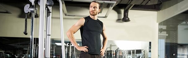 Ein athletischer Mann in aktiver Kleidung steht in einem Fitnessstudio, die Hände auf den Hüften. - Foto, Bild