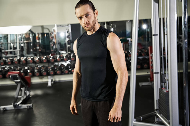 Un hombre atlético en ropa activa está de pie con confianza frente a una máquina de gimnasio, listo para entrenar y construir fuerza. - Foto, Imagen