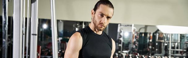 Mężczyzna w czarnej bluzce ze zbiornikiem wykonuje ćwiczenia w dobrze wyposażonej siłowni, koncentrując się na treningu siłowym i wytrzymałościowym.. - Zdjęcie, obraz