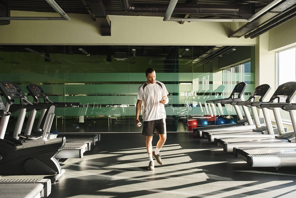 Een fitte man in atletische kleding loopt in de buurt van loopbanden in een fitnessruimte - Foto, afbeelding
