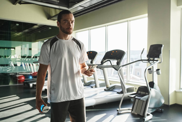 Ein athletischer Mann in aktiver Kleidung hält selbstbewusst eine Wasserflasche im Fitnessstudio. - Foto, Bild