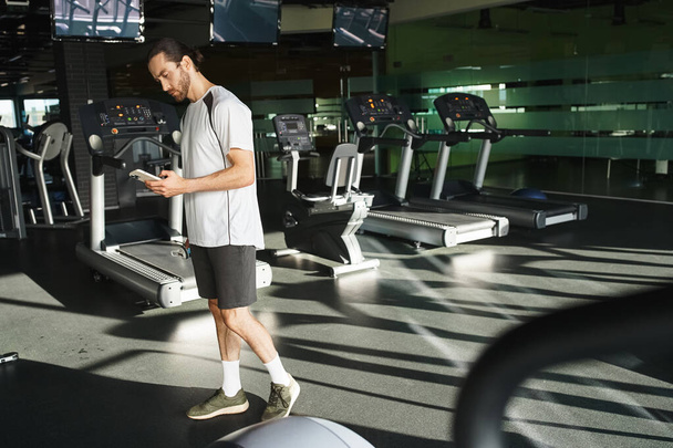 М'язистий чоловік в активному одязі перевіряє свій мобільний телефон, роблячи перерву від тренування в тренажерному залі. - Фото, зображення