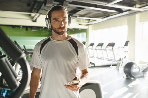 Ein athletischer Mann in aktiver Kleidung, der über Kopfhörer Musik hört und in einem lebhaften Fitnessstudio trainiert. - Foto, Bild