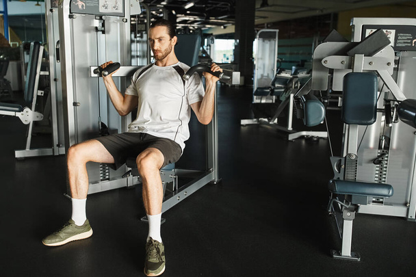 Un hombre atlético en ropa activa sentado en un banco de gimnasio, sosteniendo asas en la máquina de ejercicios - Foto, imagen
