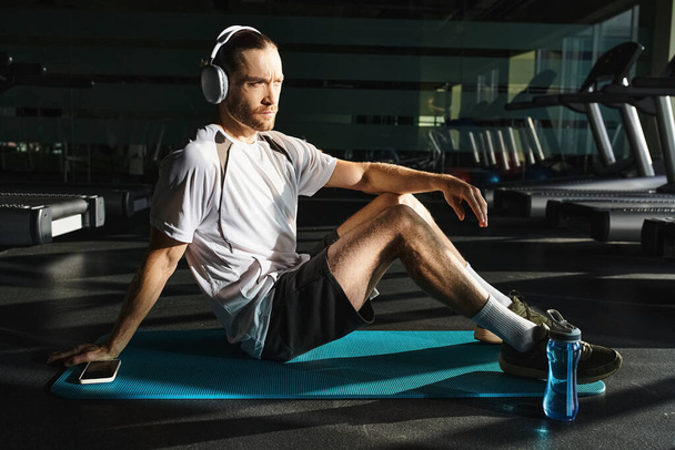 Atletický muž sedí na modrém rohožce, na uších sluchátka, ponořený do chvíle uvolnění a hudby. - Fotografie, Obrázek