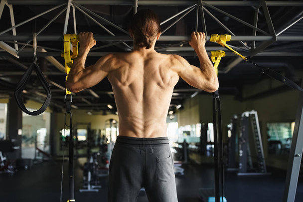 Um homem sem camisa em um ginásio executa pull ups, mostrando sua estrutura muscular e dedicação à aptidão. - Foto, Imagem
