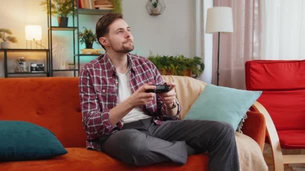 Oyun konsolu oyunu oynayan neşeli genç adam oturma odasındaki kanepede oturup eğleniyor. Beyaz adam hafta sonu evde online oyun kazanmanın keyfini çıkarıyor.. - Video, Çekim