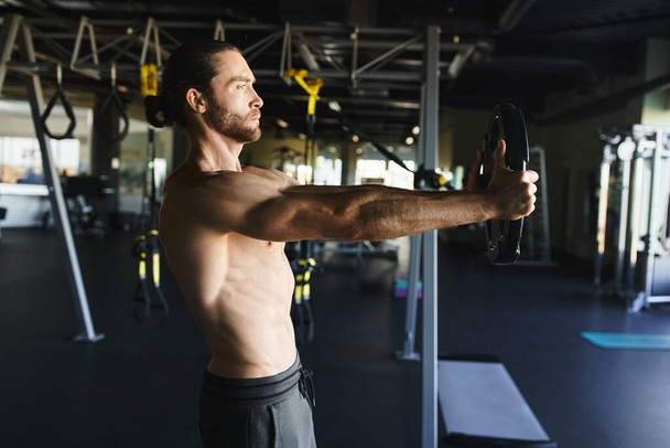 Ένας μυώδης άντρας χωρίς πουκάμισο κρατάει ένα κουδούνι σε ένα γυμναστήριο, δείχνοντας τη δύναμή του και την αφοσίωσή του στη φυσική κατάσταση.. - Φωτογραφία, εικόνα