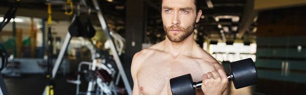Мужчина без рубашки демонстрирует свое мускулистое телосложение, держа гантель в спортзале. - Фото, изображение