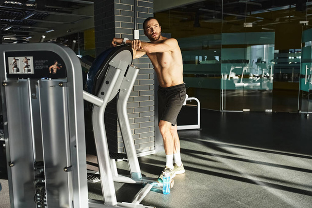Ein muskulöser Mann ohne Hemd steht selbstbewusst neben einem Trainingsgerät in einem Fitnessstudio und bereitet sich auf das Training vor.. - Foto, Bild