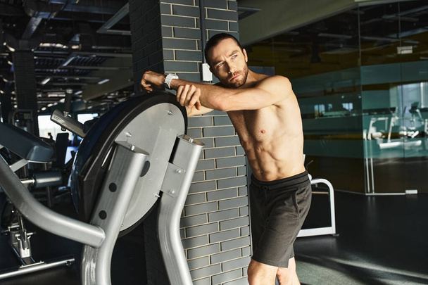 Um homem sem camisa e musculoso se inclina contra uma parede ao lado de uma máquina de exercícios em um ambiente de ginásio. - Foto, Imagem