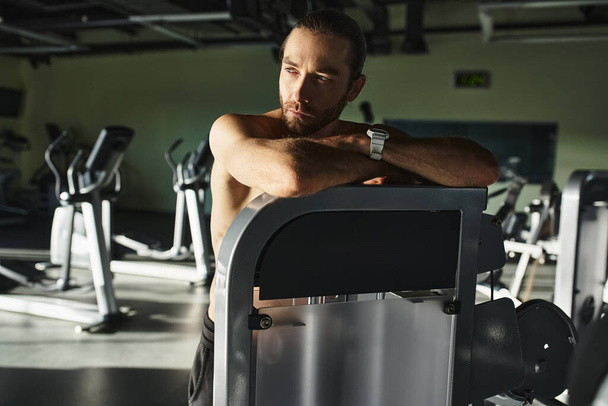 Ένας μυώδης άντρας χωρίς πουκάμισο να ακουμπά σε ένα μηχάνημα ενώ γυμνάζεται σε ένα γυμναστήριο. - Φωτογραφία, εικόνα