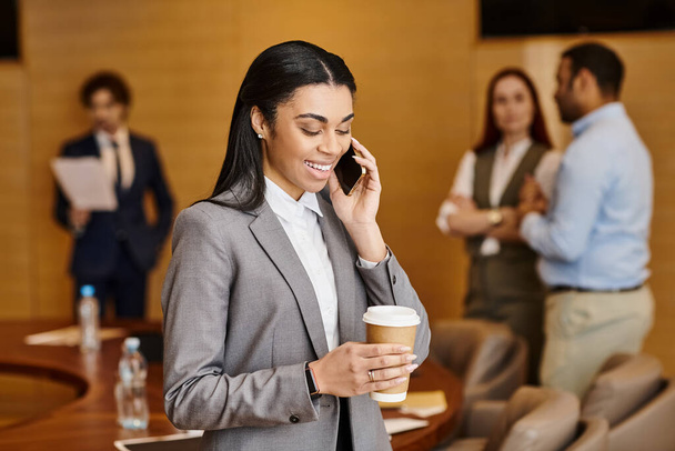 İş kıyafeti giymiş bir kadın elinde kahve tutarken cep telefonuyla konuşuyor.. - Fotoğraf, Görsel