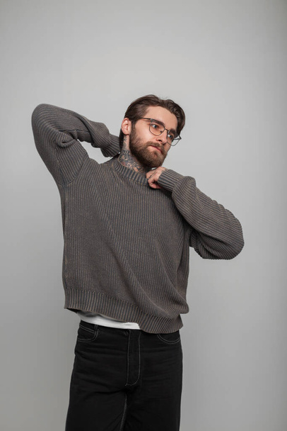 Cooler gutaussehender junger Hipster mit Frisur und Bart mit Brille in einem modisch gestrickten grauen Pullover steht und posiert auf weißem Hintergrund im Studio - Foto, Bild