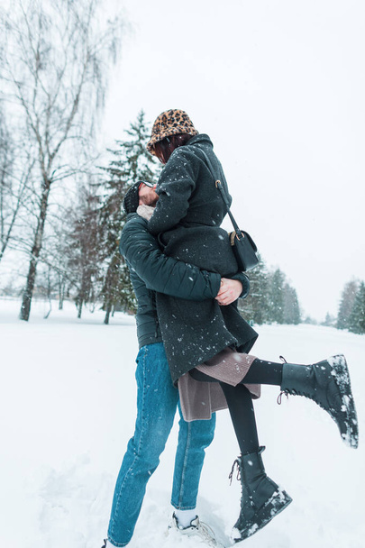 Mutlu yakışıklı adam, güzel genç kız arkadaşını karlı bir kış parkında kaldırdı. Neşeli aşıklar eğleniyor ve sarılıyorlar. - Fotoğraf, Görsel