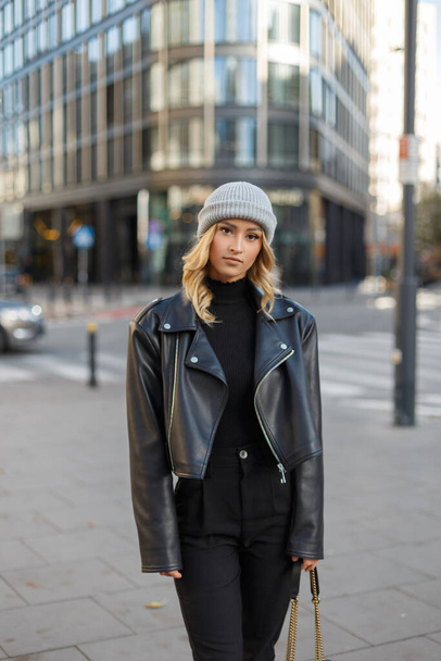 Fashion mooie urban stijlvolle meisje model met een gebreide hoed in een zwarte gebreide trui en lederen jas wandelingen in de stad - Foto, afbeelding