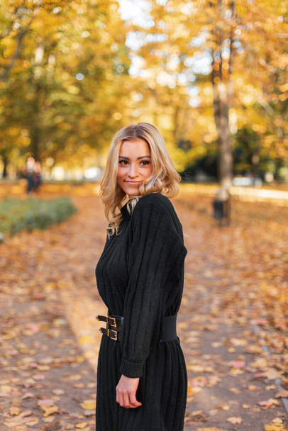 Szczęśliwy piękny młody blondynka dziewczyna w moda czarny dzianina sukienka spacery w jesiennym parku ze złotym żółtym - Zdjęcie, obraz