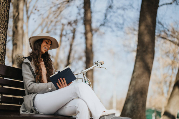 Une jeune femme à la mode profite d'un livre assis sur un banc de parc à côté de son vélo, savourant une belle journée ensoleillée. - Photo, image