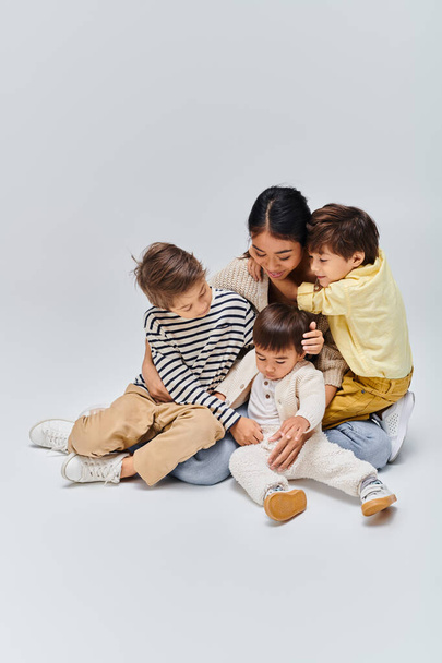 若いアジアの母親がグレーのスタジオの背景に落ち着いた穏やかな瞬間に子供たちと地面に座る. - 写真・画像