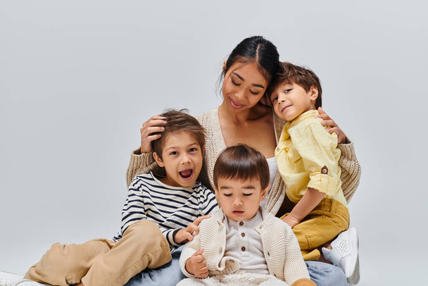 Una giovane madre asiatica si siede su un letto, circondata dai suoi figli, condividendo un momento di pace e amore in un ambiente da studio. - Foto, immagini