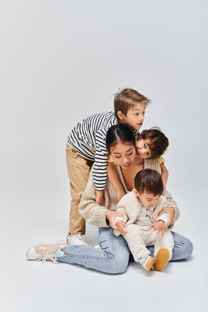 Młoda azjatycka matka i jej dzieci siedzą na sobie w studio na szarym tle. - Zdjęcie, obraz