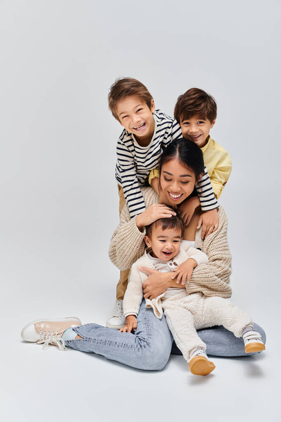 若いアジアの母親が床に座り,子供たちが灰色の背景を背景にスタジオで座っています.. - 写真・画像