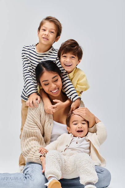 Eine junge asiatische Mutter sitzt mit ihren Kindern auf dem Boden und umarmt sich in einem Studio vor grauem Hintergrund liebevoll. - Foto, Bild