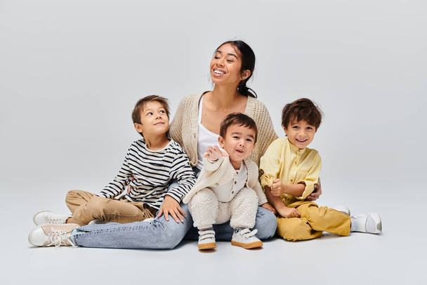 A young Asian mother sits on the ground with her children beside her in a studio setting on a grey background. - Photo, Image