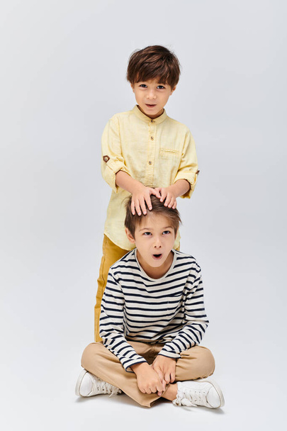 Двоє молодих хлопчиків сидять на землі, руки над головою, заблукали в роздумах на сірому студійному фоні - Фото, зображення