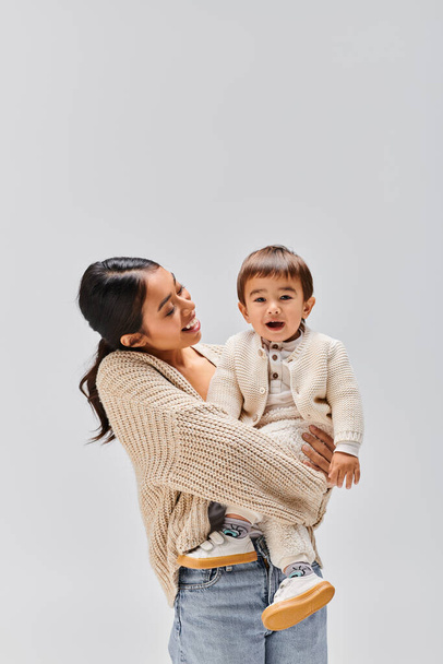 Μια νεαρή Ασιάτισσα μητέρα να κρατάει το μωρό της στην αγκαλιά της σε ένα στούντιο σε γκρι φόντο.. - Φωτογραφία, εικόνα