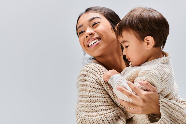 Una giovane madre asiatica culla il suo bambino in un tenero abbraccio su uno sfondo grigio. - Foto, immagini