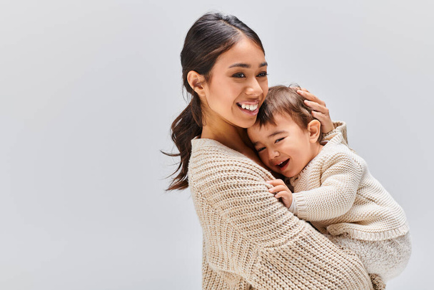 Eine junge asiatische Mutter umarmt ihr Kind zärtlich in einem Studio vor grauem Hintergrund. - Foto, Bild