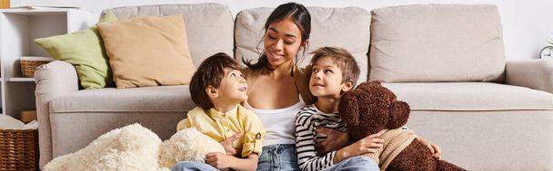 Genç bir Asyalı anne, sıcak ve sevgi dolu bir ilişki içinde, sıcak bir oturma odasında iki çocuğuyla bir kanepede oturuyor.. - Fotoğraf, Görsel