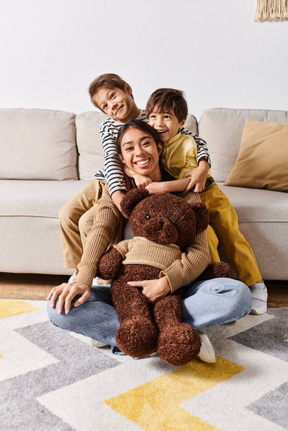 Een jonge Aziatische moeder zit op de vloer met haar twee zoontjes en een teddybeer, genietend van een vredig moment samen. - Foto, afbeelding
