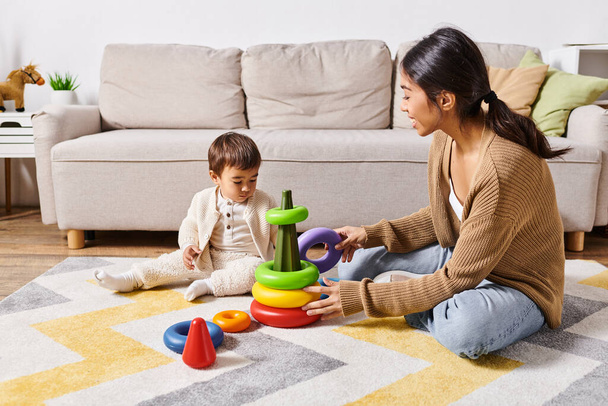 Młoda azjatycka matka radośnie współpracuje ze swoim synkiem, bawiąc się razem na podłodze w salonie. - Zdjęcie, obraz