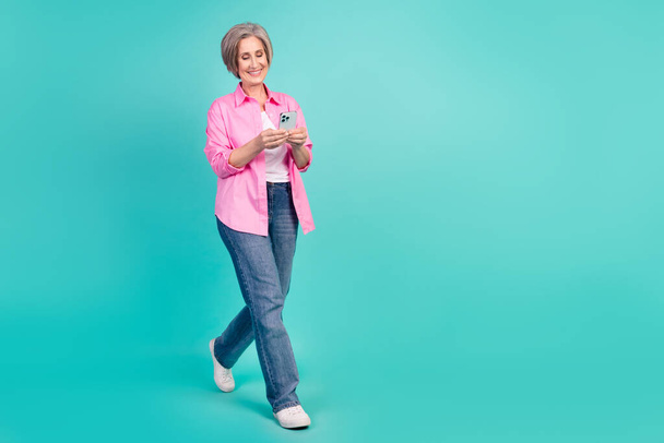 Full length photo of smart retired woman wear pink shirt read email on smartphone go to empty space isolated on teal color background. - Фото, изображение