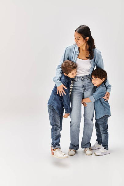 Eine junge asiatische Mutter und ihre kleinen Söhne, alle in Jeanskleidung, posieren in einem grauen Studio für ein Foto. - Foto, Bild