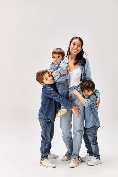 Una joven madre asiática posando con sus hijos pequeños, todos vestidos de mezclilla, capturando un momento conmovedor en un estudio gris. - Foto, imagen