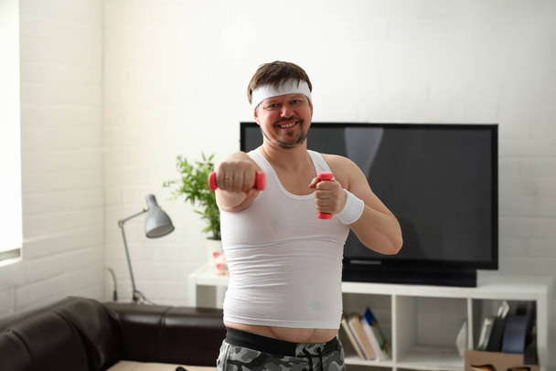 junger attraktiver Mann, der zu Hause Fitness betreibt und eine Hantel in der Hand hält, um Übergewicht und Verlust zu bekämpfen - Foto, Bild