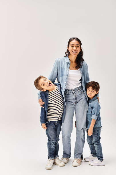 Eine junge asiatische Mutter und ihre kleinen Söhne, alle in Jeans-Outfits, posieren gemeinsam für ein Porträt in einem grauen Studio. - Foto, Bild
