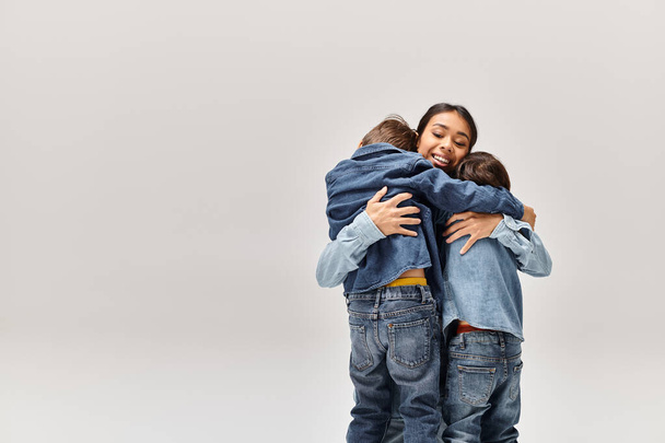 Молодая азиатская мать и ее маленькие сыновья, одетые в джинсовую одежду, обнимают друг друга на белом фоне. - Фото, изображение
